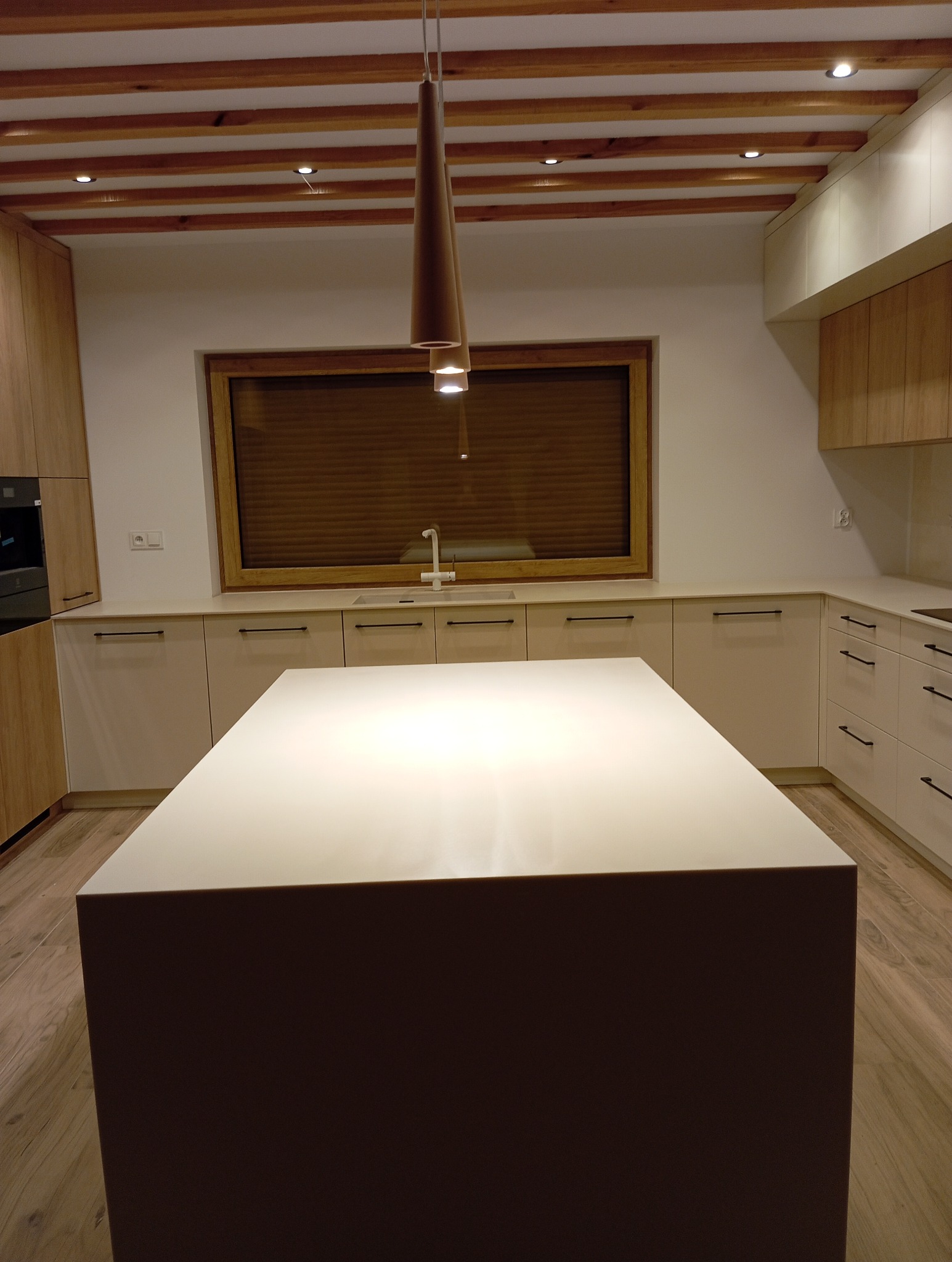 Nowoczesna kuchnia z drewnianymi akcentami: podwieszane oświetlenie nad wyspą kuchenną, drewniane belki na suficie i zabudowa meblowa z elementami drewna.