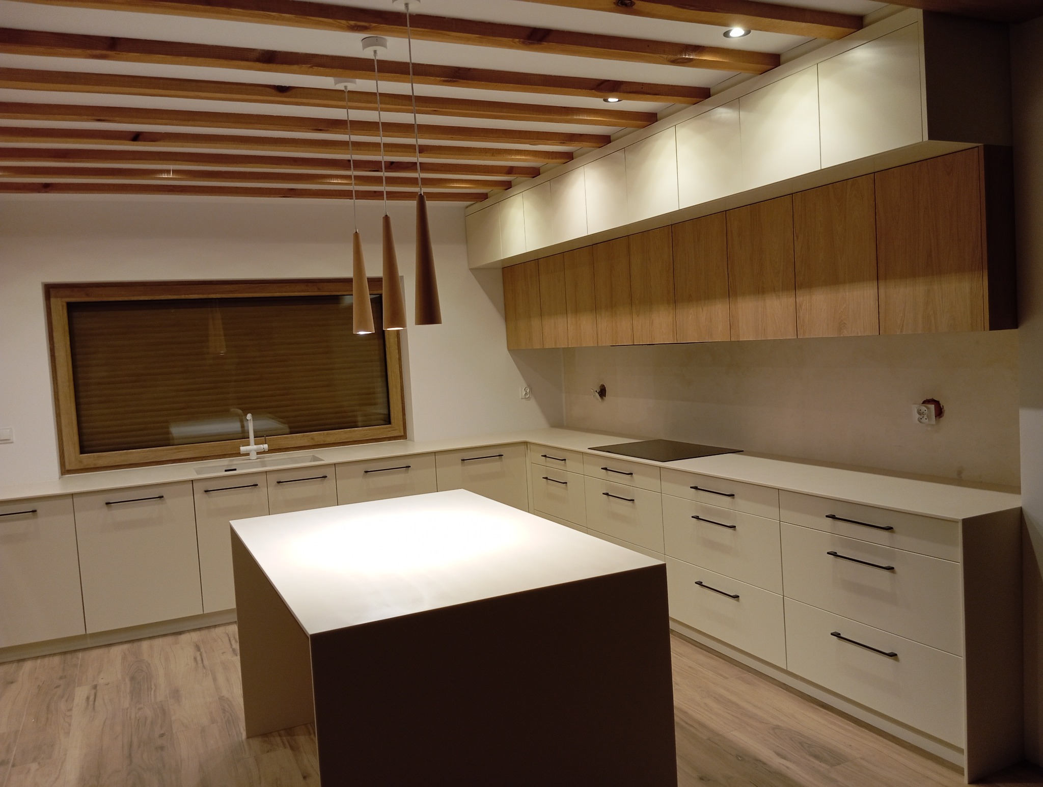 Nowoczesna, jasna kuchnia z drewnianymi akcentami: wyspa, szafki wiszące z drewnianymi frontami, drewniane belki na suficie i trzy wiszące lampy o stożkowym kształcie.