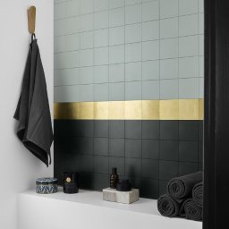 Centrum Napraw Domowych - Ściana łazienki wyłożona kwadratowymi płytkami w kolorze pastelowej zieleni i czerni, oddzielona poziomym pasem złotych płytek, z ręcznikiem na drewnianym wieszaku, kosmetykami i zwiniętymi...