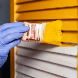 Malowanie drewnianych żaluzji na żółto pędzlem trzymanym w dłoni w niebieskiej rękawicy ochronnej. Widoczne ślady świeżej farby.