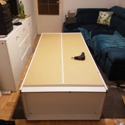 Marcel Durczok - Płyta meblowa z białymi obrzeżami na podłodze w trakcie montażu, w tle wkrętarka akumulatorowa, parkiet i fragment sofy.