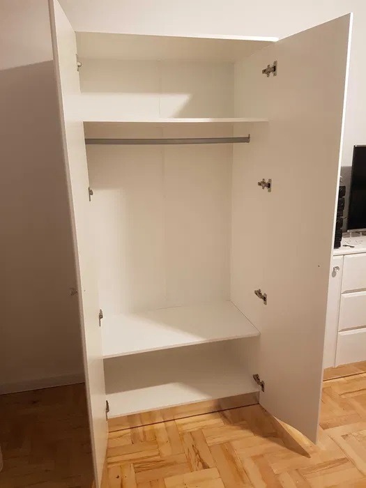 Wnętrze białej, otwartej szafy z półkami i drążkiem na ubrania, widoczne zawiasy, podłoga parkietowa w tle.