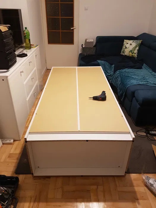 Płyta meblowa z białymi obrzeżami na podłodze w trakcie montażu, w tle wkrętarka akumulatorowa, parkiet i fragment sofy.