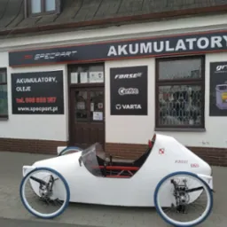 Sklep Specpart z akumulatorami i olejami, przed budynkiem biały pojazd trójkołowy z niebieskimi oponami, z nazwą Piskorz IKO.