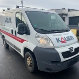 Biały bus Peugeot z logo firmy JER i KAUER na karoserii, zaparkowany na mokrym asfalcie przed nowoczesnym domem.