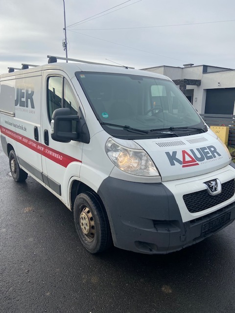 Biały bus Peugeot z logo firmy JER i KAUER na karoserii, zaparkowany na mokrym asfalcie przed nowoczesnym domem.