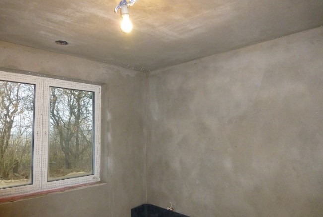 Wnętrze pokoju po nałożeniu świeżego, jasnoszarego tynku, widoczne okno z białymi ramami i widokiem na drzewa bez liści, surowa żarówka zwisająca z sufitu.