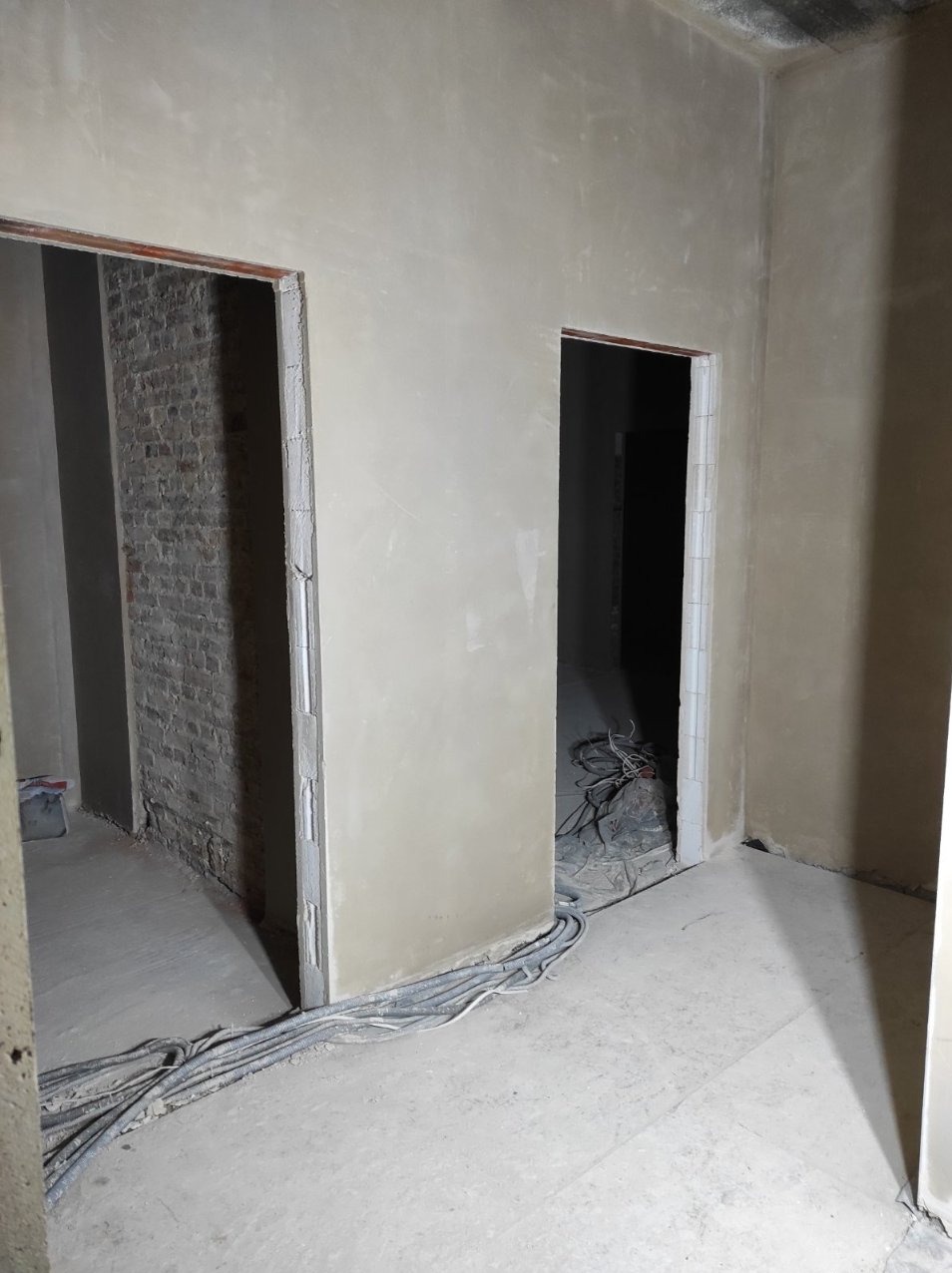 Wnętrze w trakcie prac wykończeniowych: ściany pokryte świeżym, jasnym tynkiem, widoczne otwory drzwiowe, w jednym z nich surowa cegła, na podłodze kable instalacji elektrycznej.