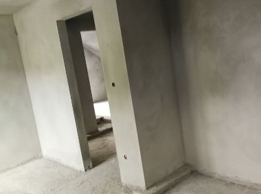 Surowe wnętrze w trakcie prac wykończeniowych, widoczne świeżo otynkowane ściany w szarym odcieniu, z widocznymi otworami pod gniazdka elektryczne i fragmentem podłogi pokrytej resztkami zaprawy.