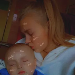 Blondwłosa kobieta całuje w czoło śpiącego chłopca w niebieskiej koszulce, oboje z filtrem 'Babe' na twarzach.