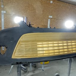 L&M DESIGN - Przygotowany do lakierowania zderzak samochodowy, zabezpieczony taśmą maskującą, na metalowym stojaku w warsztacie.