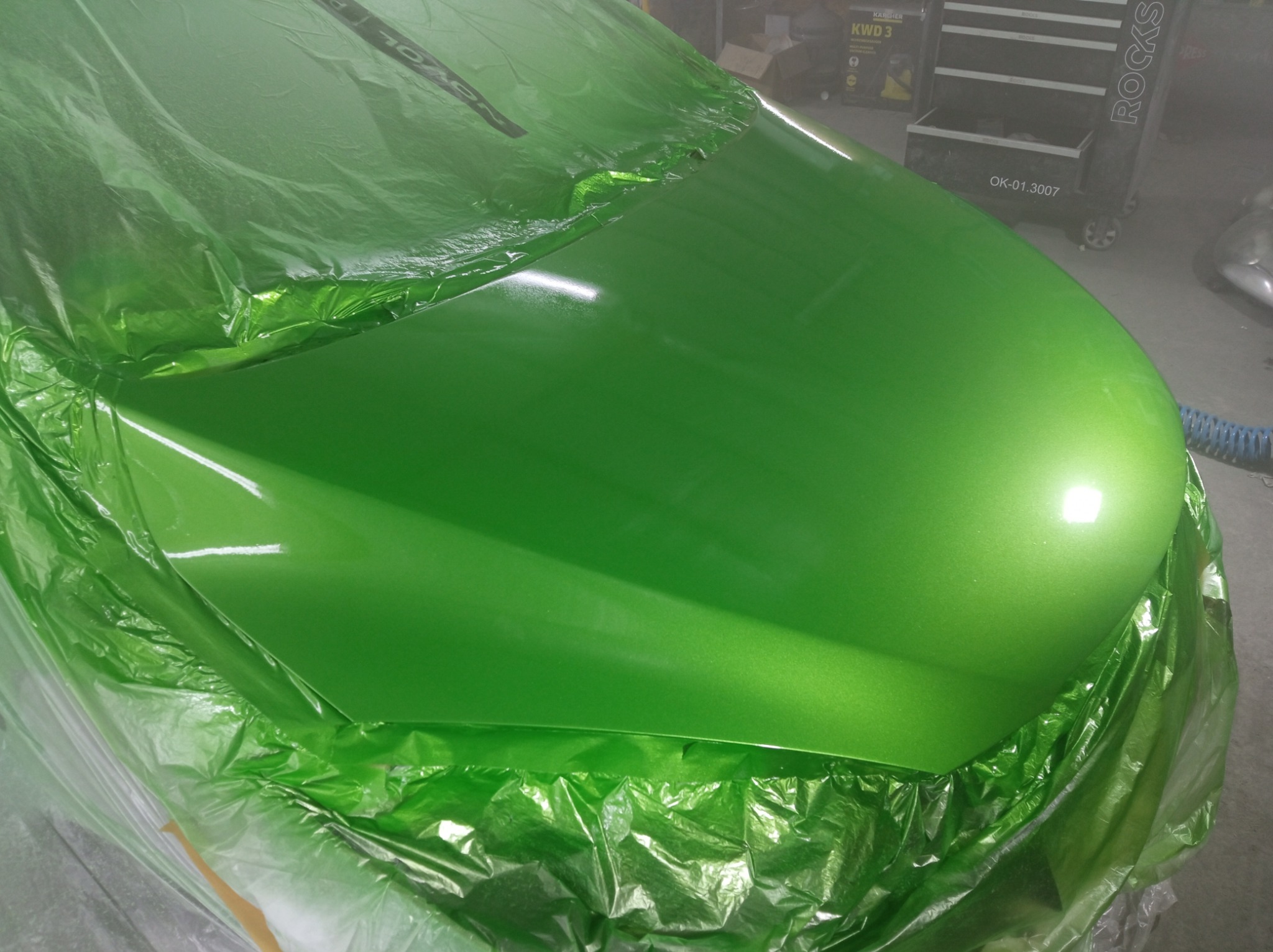 Świeżo polakierowana, zielona maska samochodu w Siewierzu, zabezpieczona folią ochronną. Widoczne odbicie światła na gładkiej powierzchni lakieru, z tyłu w tle wózek warsztatowy.