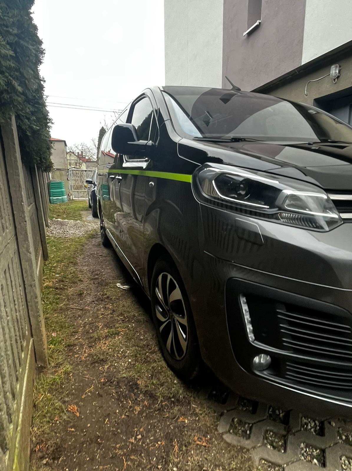 Szary samochód dostawczy z neonowo-zielonym paskiem bocznym zaparkowany na nierównej nawierzchni z kostki brukowej, widok z boku na tle ogrodzenia i budynku.