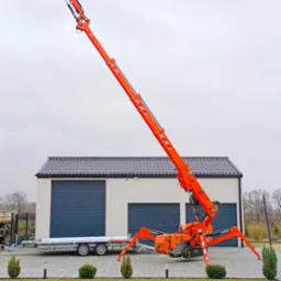 Pomarańczowy pająk-dźwig ustawiony na kostce brukowej przed nowoczesnym garażem z dwiema bramami rolowanymi, wysięgnik skierowany w górę, podczepiona przyczepa.