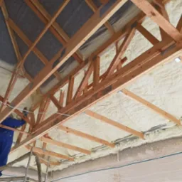 Pracownik w niebieskim kombinezonie aplikuje pianę izolacyjną na poddasze, widoczne drewniane krokwie i częściowo pokryta pianą przestrzeń pod dachem.