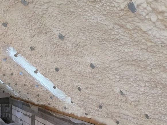 Poddasze po natrysku pianki izolacyjnej, widoczne szare punkty mocowania i biała linia uszczelnienia, surowa konstrukcja z cegieł i drewna w dolnej części kadru.