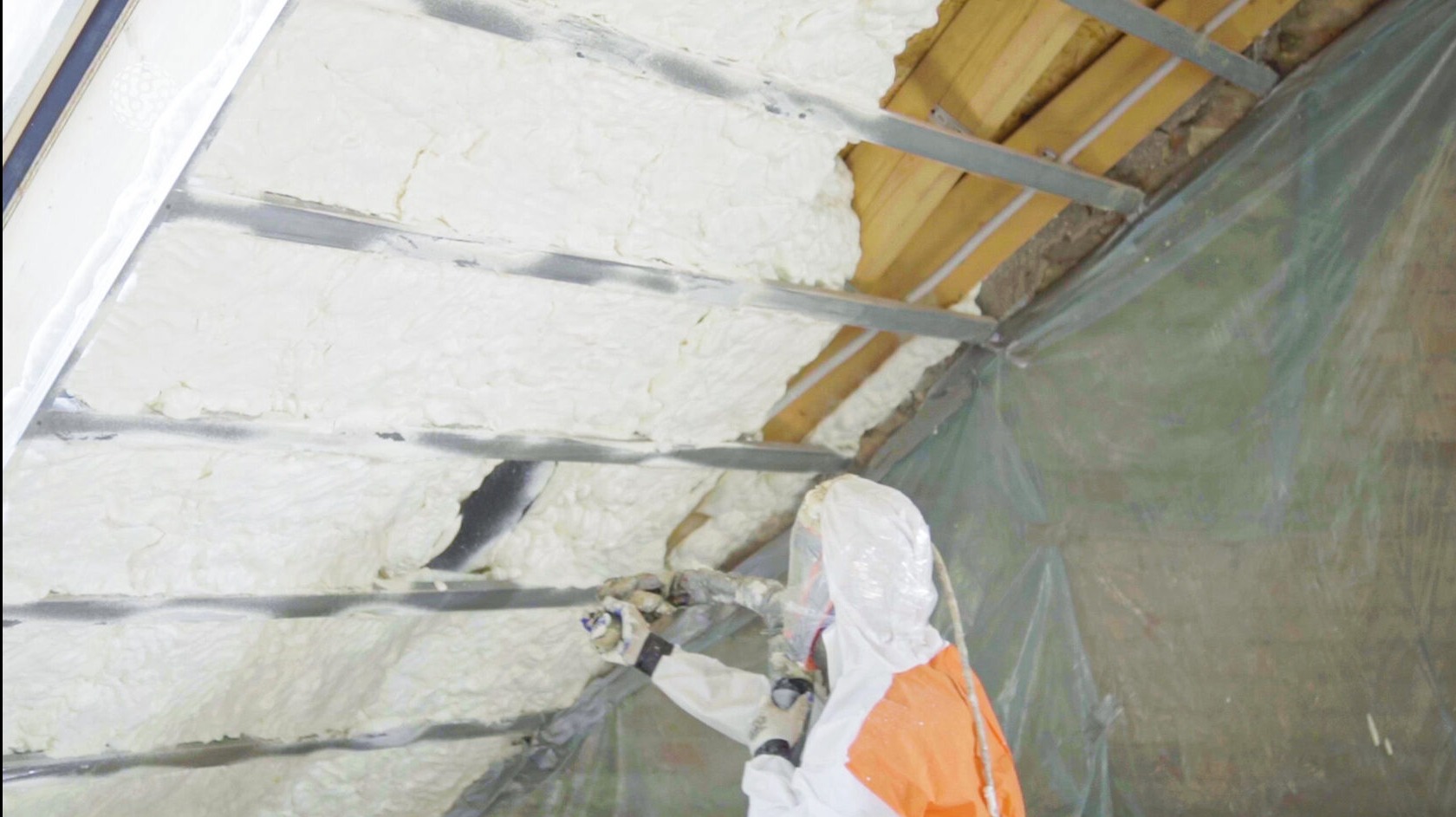 Pracownik w kombinezonie ochronnym aplikuje pianę izolacyjną na poddaszu między metalowymi profilami, widoczne drewniane elementy konstrukcji dachu i folia ochronna.