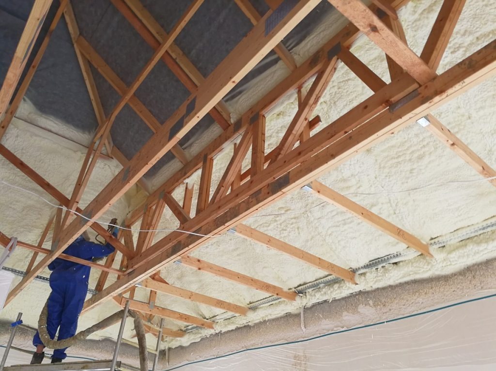 Pracownik w niebieskim kombinezonie aplikuje pianę izolacyjną na poddasze, widoczne drewniane krokwie i częściowo pokryta pianą przestrzeń pod dachem.