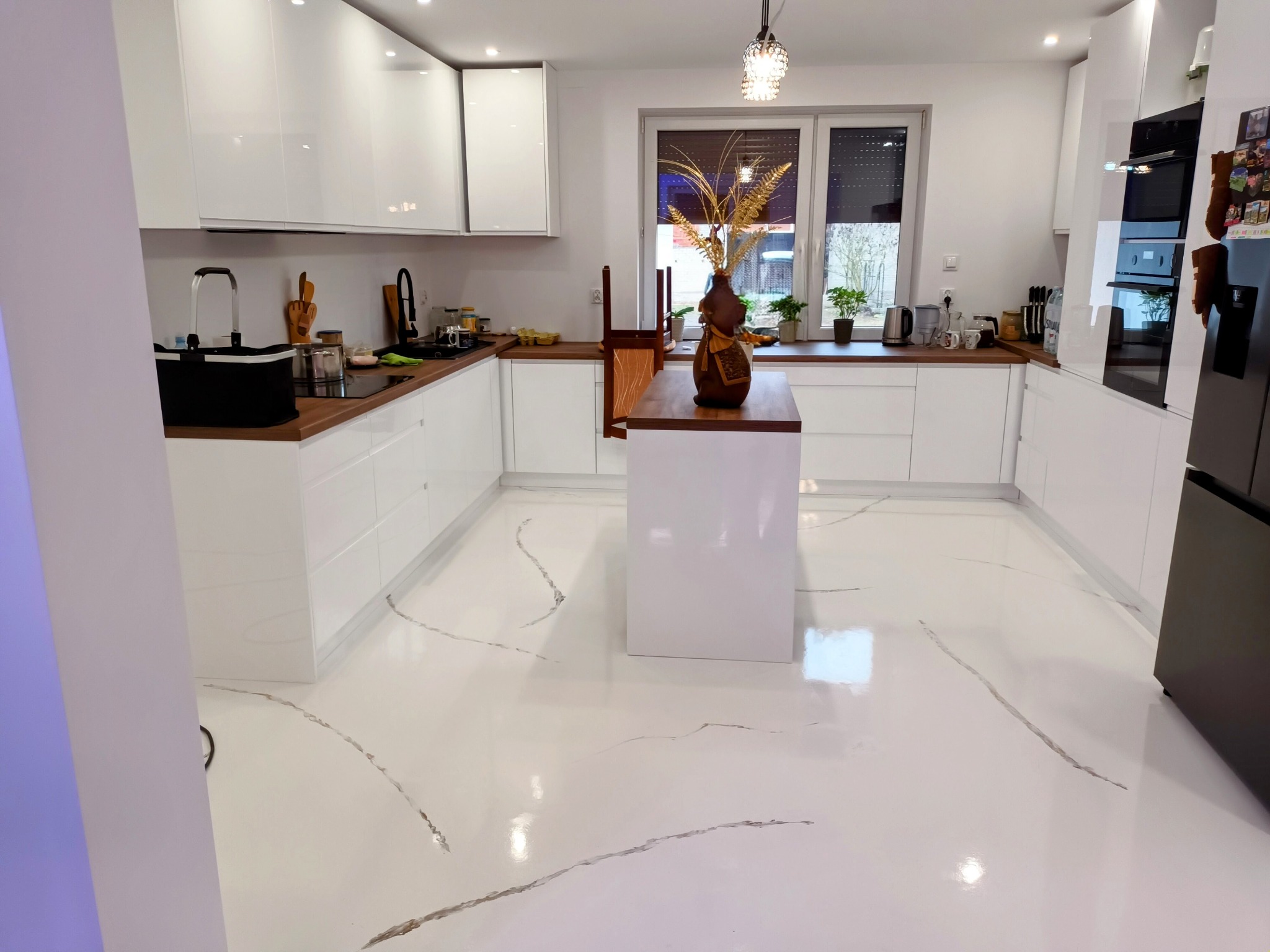 Biała kuchnia z wyspą i błyszczącą podłogą z szarymi żyłkami imitującymi marmur.