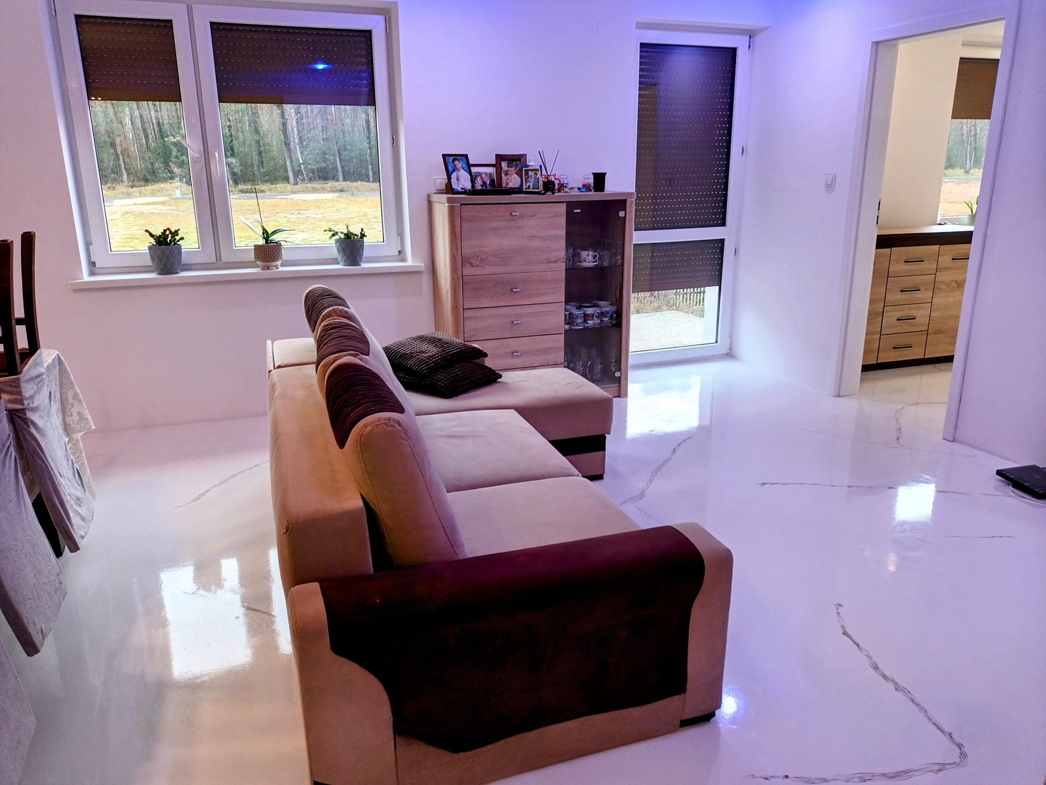 Biała, błyszcząca podłoga z delikatnymi szarymi żyłkami w salonie, widok z perspektywy sofy w kierunku okna i otwartego przejścia do kuchni.