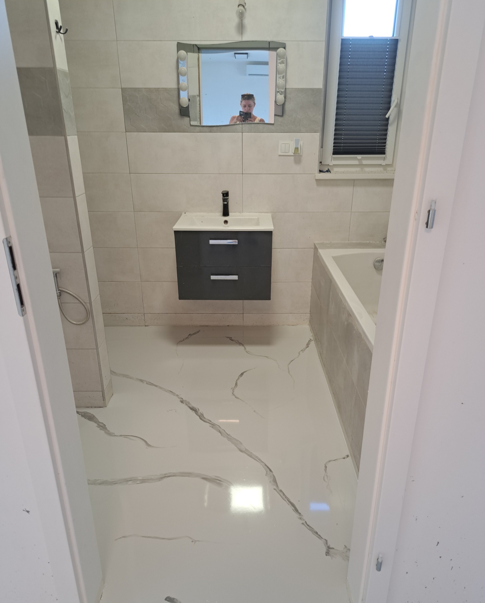 Biała posadzka w łazience z szarymi żyłkami imitującymi marmur, widoczna umywalka z szafką, wanna i fragment kabiny prysznicowej.