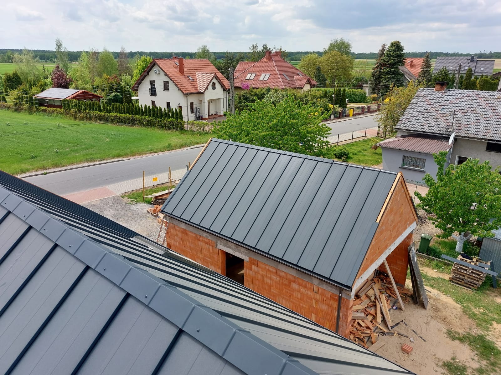 Widok z góry na nowo kryty blachą dachówkową garaż z cegły, z widocznymi elementami konstrukcyjnymi, na tle domów jednorodzinnych i zieleni.