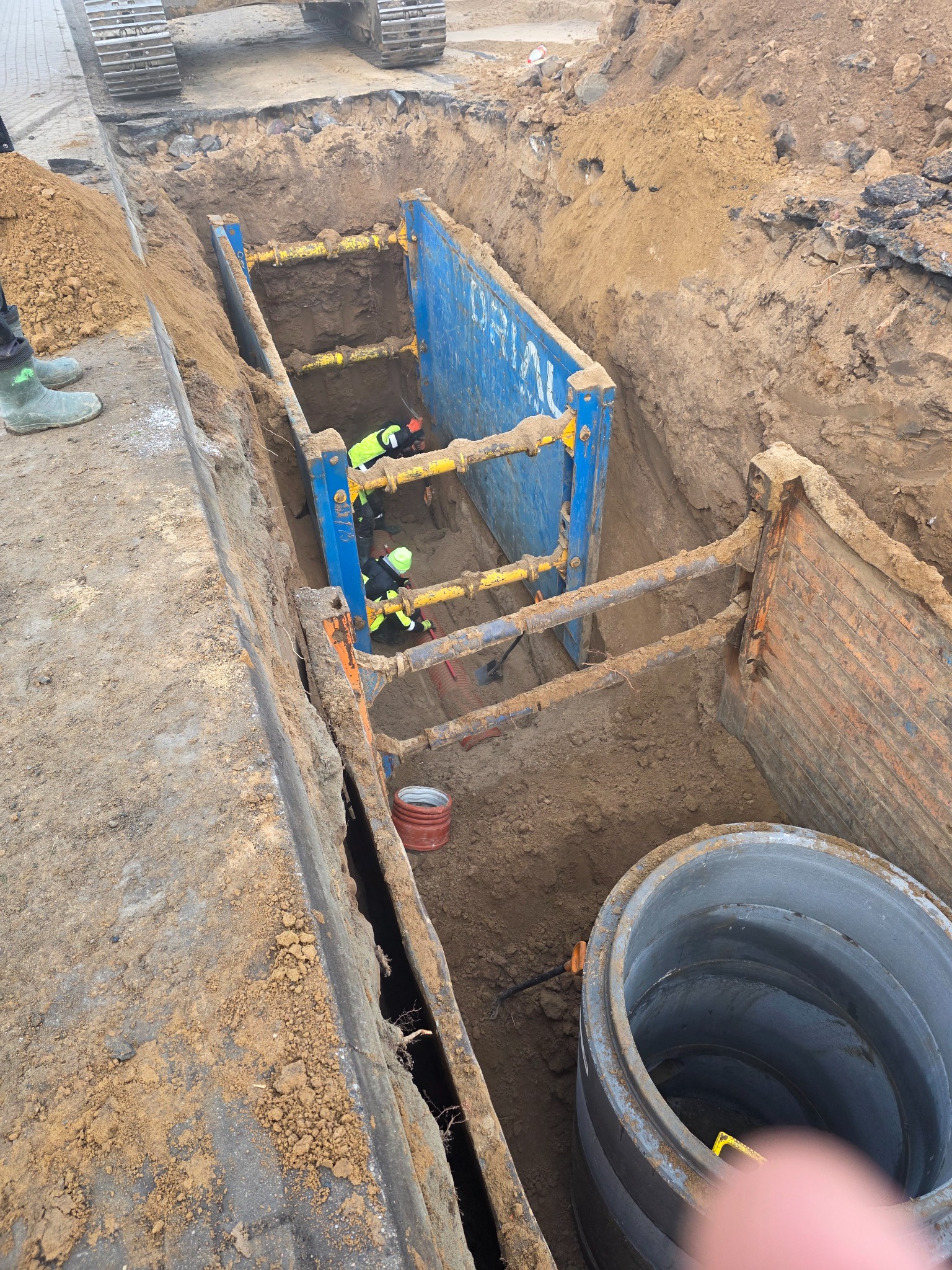 Głęboki wykop zabezpieczony szalunkiem stalowym, wewnątrz pracownicy w odblaskowych ubraniach. Na pierwszym planie betonowy krąg studzienny i fragment palca.