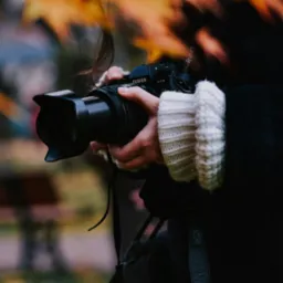 Osoba trzyma aparat Fujifilm X-T3 z założonym obiektywem, jesienne liście w tle.