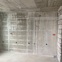 EL-PRO Instalacje Elektryczne i Teletechniczne - Surowy betonowy pokój w trakcie instalacji elektrycznej: białe kable prowadzone po ścianach i suficie, widoczne otwory na gniazdka, pomarańczowe puszki instalacyjne i fragment murowanej kolumny...