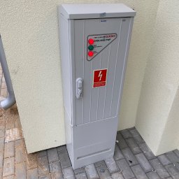 EL-PRO Instalacje Elektryczne i Teletechniczne - Szara szafka elektryczna na zewnątrz budynku z sygnalizacją LED (czerwona i zielona) i ostrzeżeniem przeciwpożarowym. Widoczne oznaczenia producenta Pelmet i Cerbex.