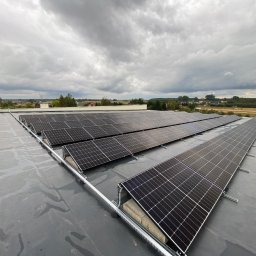 EL-PRO Instalacje Elektryczne i Teletechniczne - Rząd paneli słonecznych zamontowanych na płaskim, szarym dachu, z widokiem na krajobraz pod pochmurnym niebem.