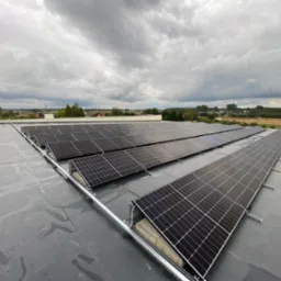 Rząd paneli słonecznych zamontowanych na płaskim, szarym dachu, z widokiem na krajobraz pod pochmurnym niebem.
