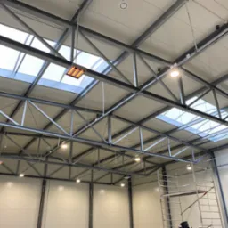 Wysoki sufit hali przemysłowej z widocznymi elementami konstrukcyjnymi, oświetleniem LED i grzejnikiem przemysłowym pod oknem dachowym, z rusztowaniem w rogu.