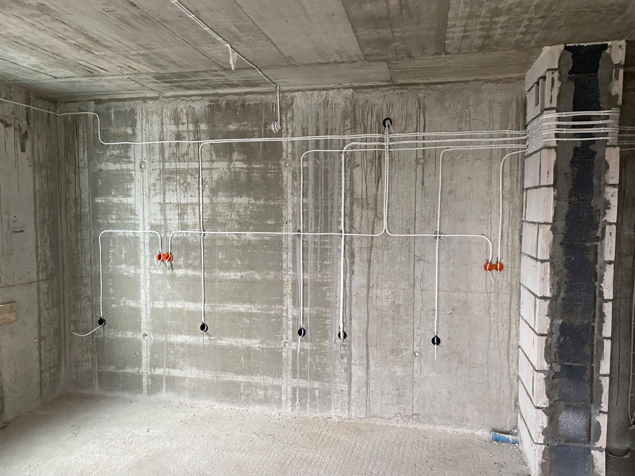 Surowy betonowy pokój w trakcie instalacji elektrycznej: białe kable prowadzone po ścianach i suficie, widoczne otwory na gniazdka, pomarańczowe puszki instalacyjne i fragment murowanej kolumny...