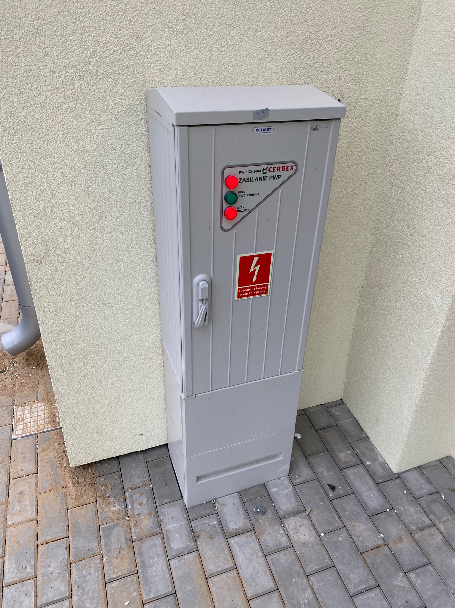 Szara szafka elektryczna na zewnątrz budynku z sygnalizacją LED (czerwona i zielona) i ostrzeżeniem przeciwpożarowym. Widoczne oznaczenia producenta Pelmet i Cerbex.