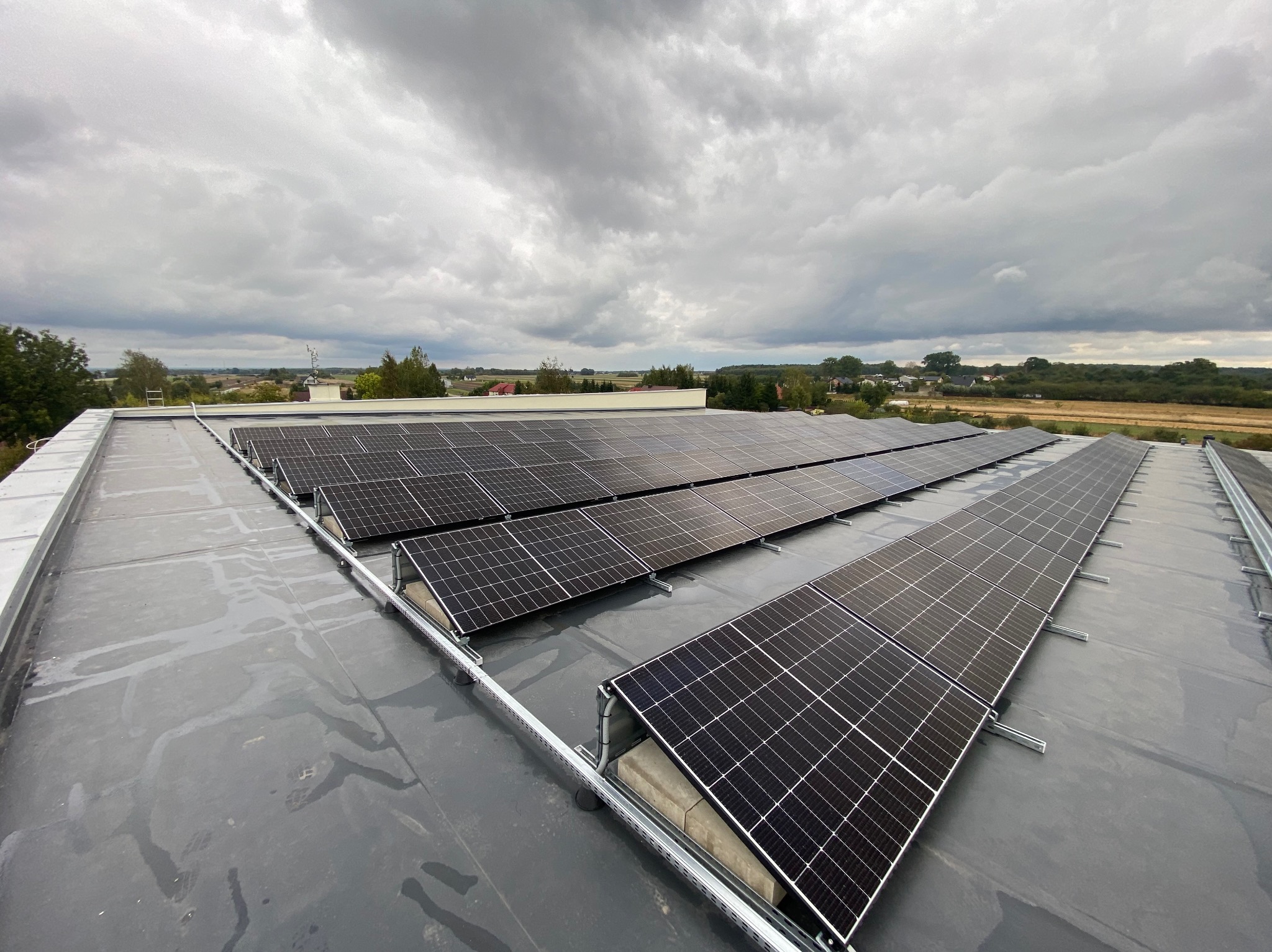 Rząd paneli słonecznych zamontowanych na płaskim, szarym dachu, z widokiem na krajobraz pod pochmurnym niebem.