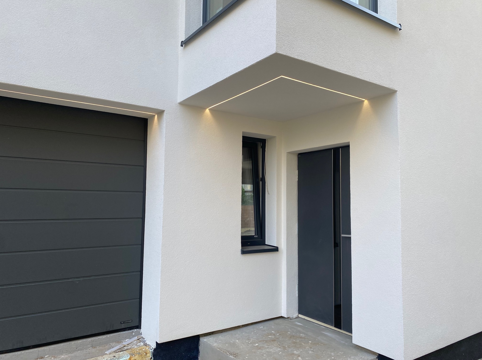 Nowoczesna fasada budynku z oświetleniem LED pod okapem i nad bramą garażową, grafitowa brama segmentowa i drzwi wejściowe, biały tynk strukturalny.