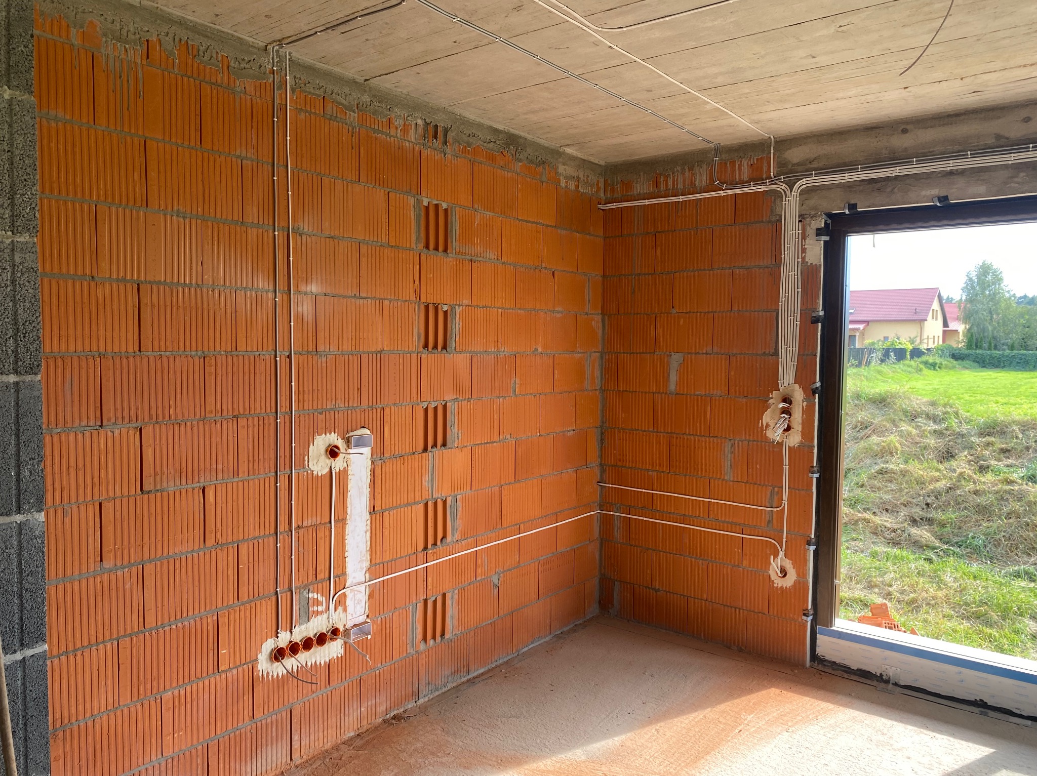 Wnętrze surowego budynku z widoczną instalacją elektryczną w peszlach, prowadzoną po ceglanych ścianach i betonowym suficie, z puszkami instalacyjnymi pod gniazdka i włączniki, oraz widokiem...