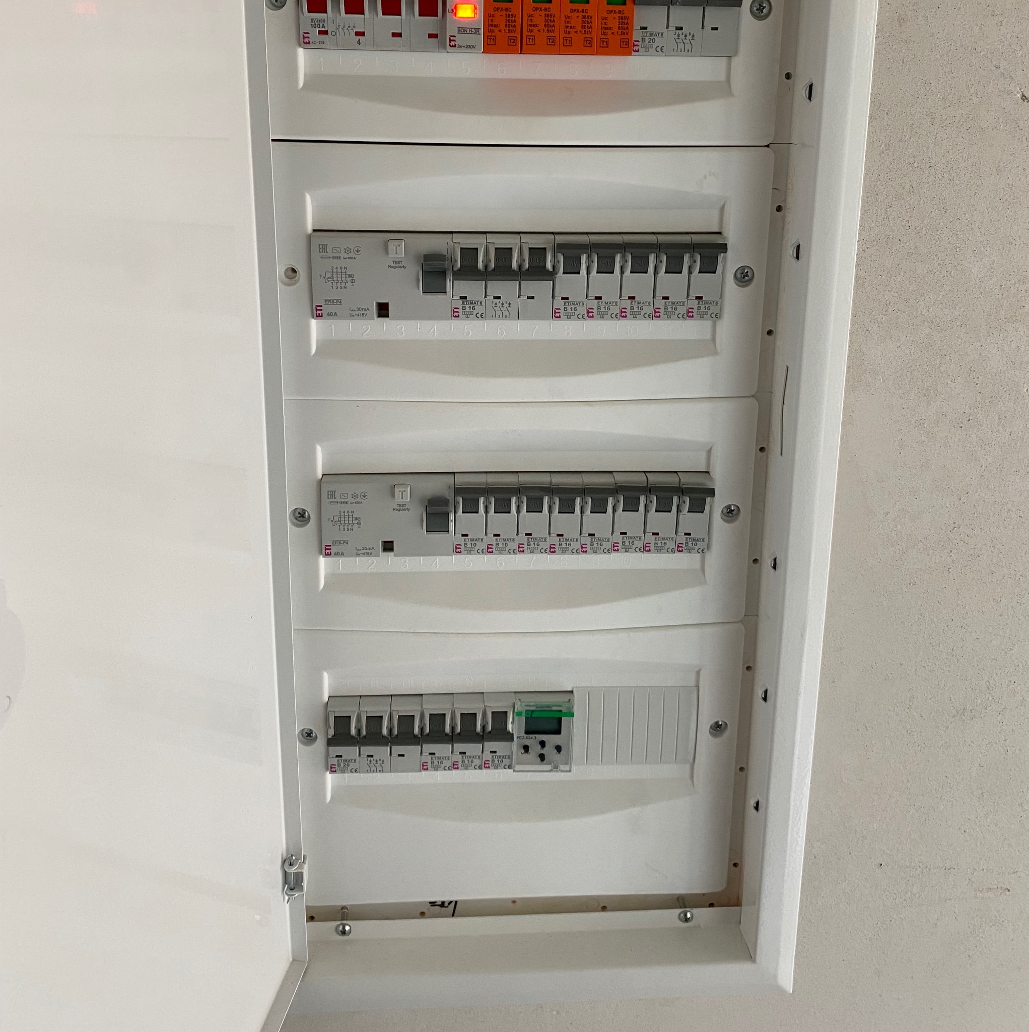Otwarta skrzynka rozdzielcza z bezpiecznikami i wyłącznikami różnicowoprądowymi marki ETI, zamontowana na szarej ścianie, widoczne numery obwodów.