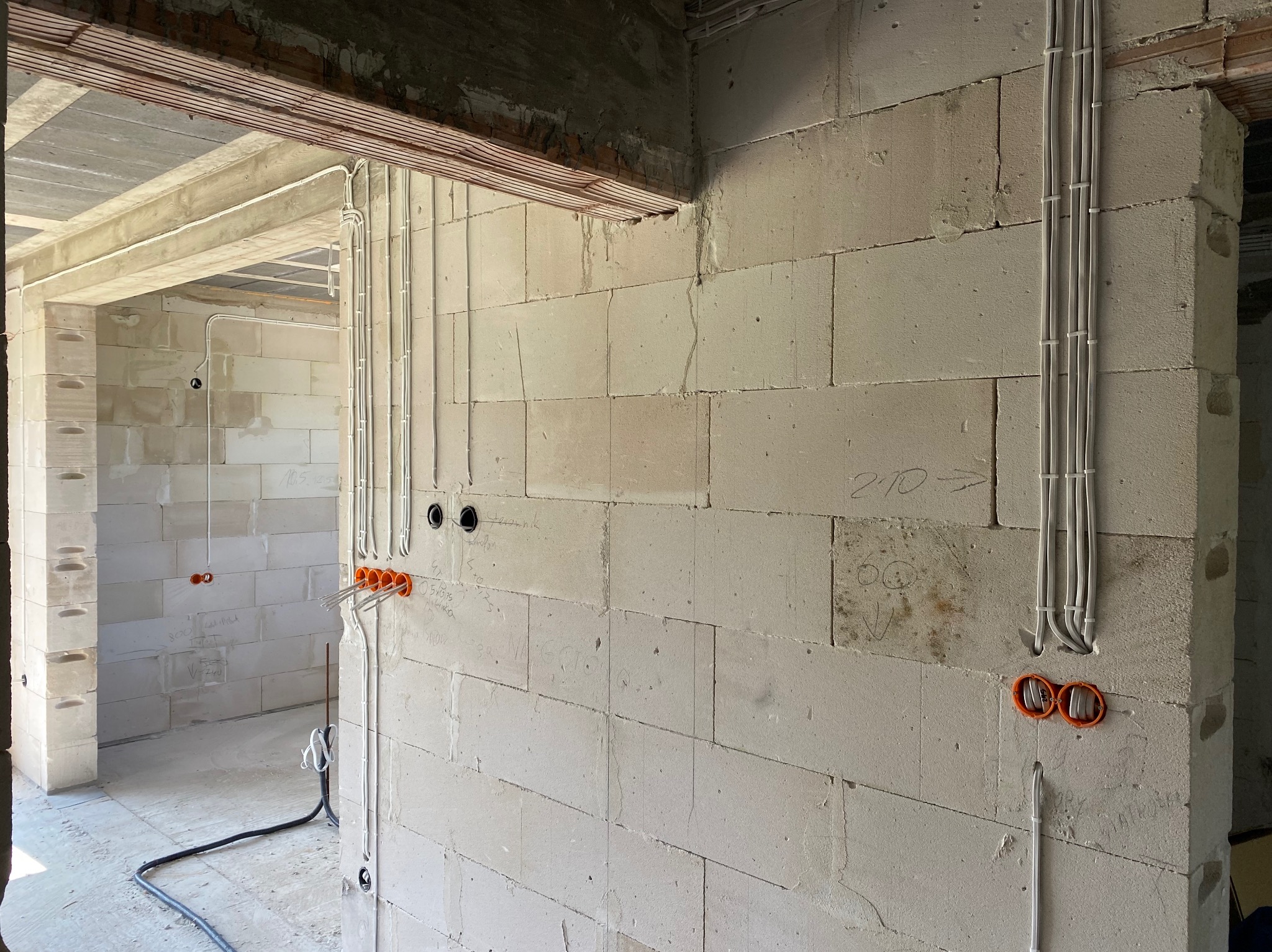 Surowe wnętrze w trakcie prac: widoczne białe ściany z betonu komórkowego, kable elektryczne prowadzone natynkowo z pomarańczowymi puszkami instalacyjnymi, oświetlenie tymczasowe.