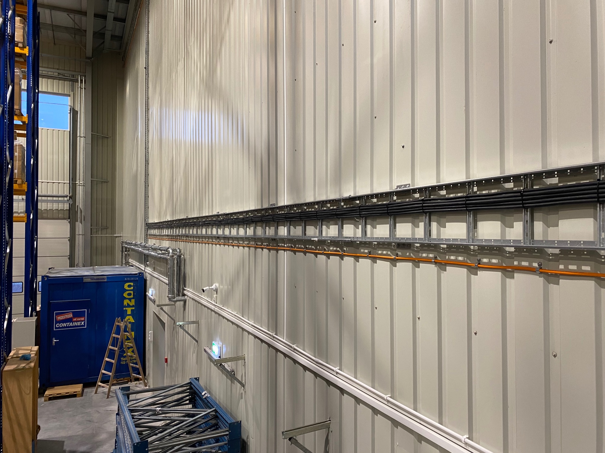 Wnętrze hali magazynowej z widocznymi trasami kablowymi prowadzonymi w korytach metalowych i rurkach PCV, oraz zamontowaną kamerą przemysłową na ścianie z blachy trapezowej.