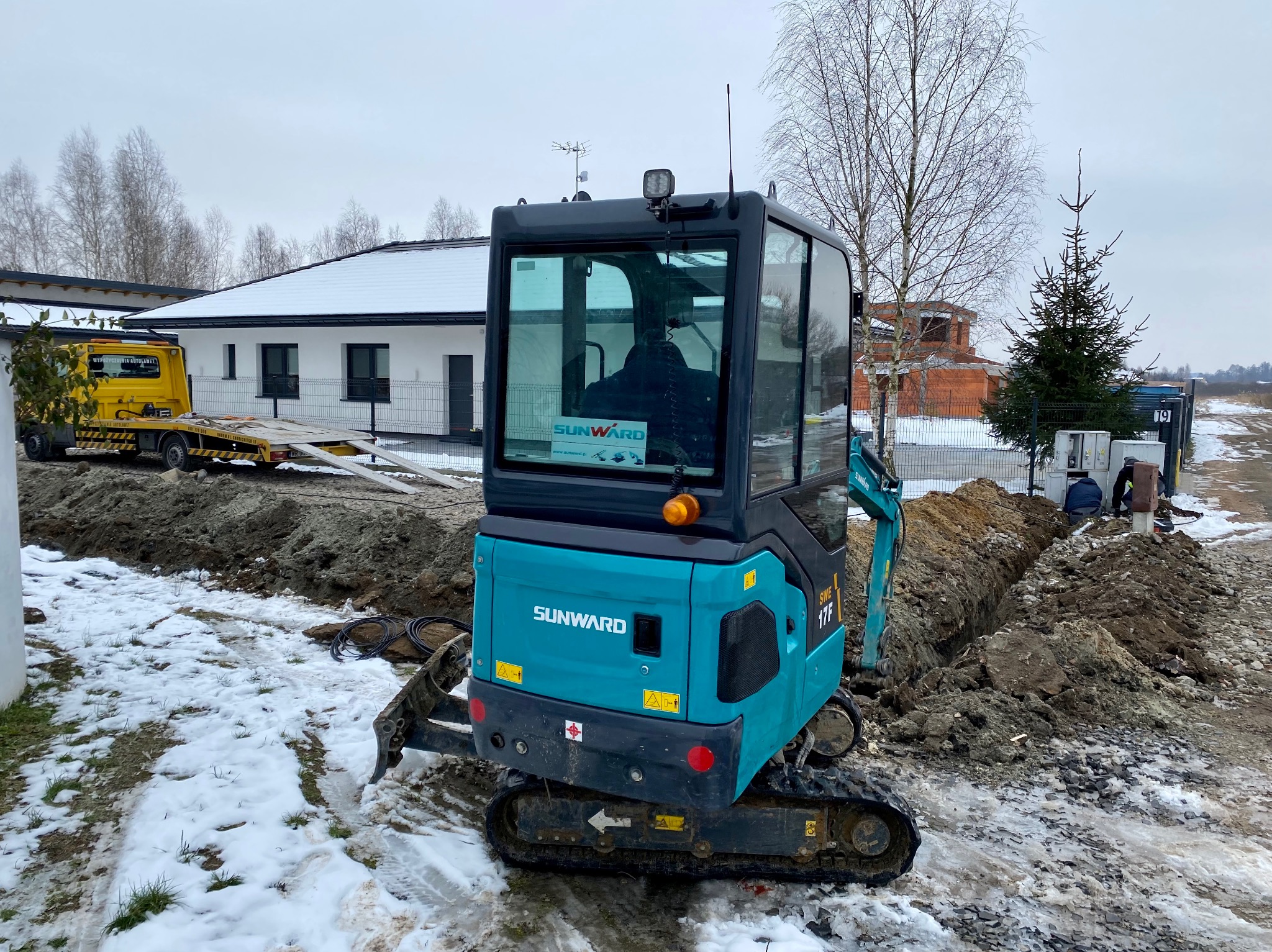 Niebiesko-czarna mini koparka Sunward SWE 17F na placu budowy z ziemią i śniegiem, w tle dom i laweta z żółtą ciężarówką.