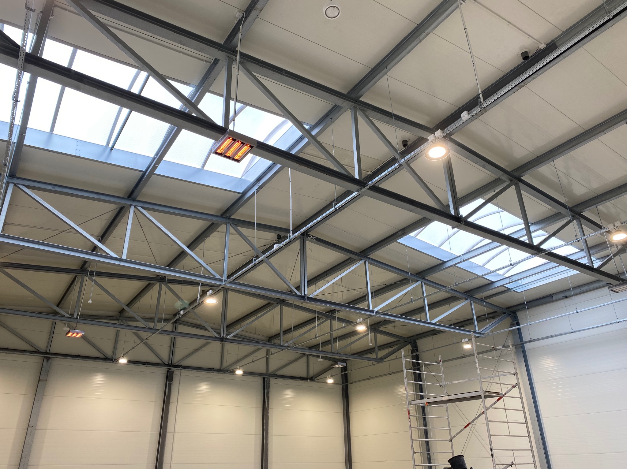Wysoki sufit hali przemysłowej z widocznymi elementami konstrukcyjnymi, oświetleniem LED i grzejnikiem przemysłowym pod oknem dachowym, z rusztowaniem w rogu.
