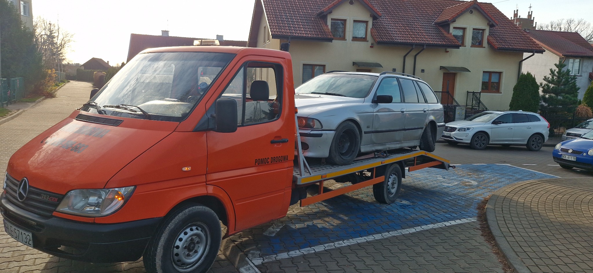 Pomarańczowa laweta Mercedes-Benz Sprinter z załadowanym srebrnym kombi BMW E39 na tle domów jednorodzinnych z czerwonymi dachami, zaparkowana na parkingu z niebieską kostką brukową.