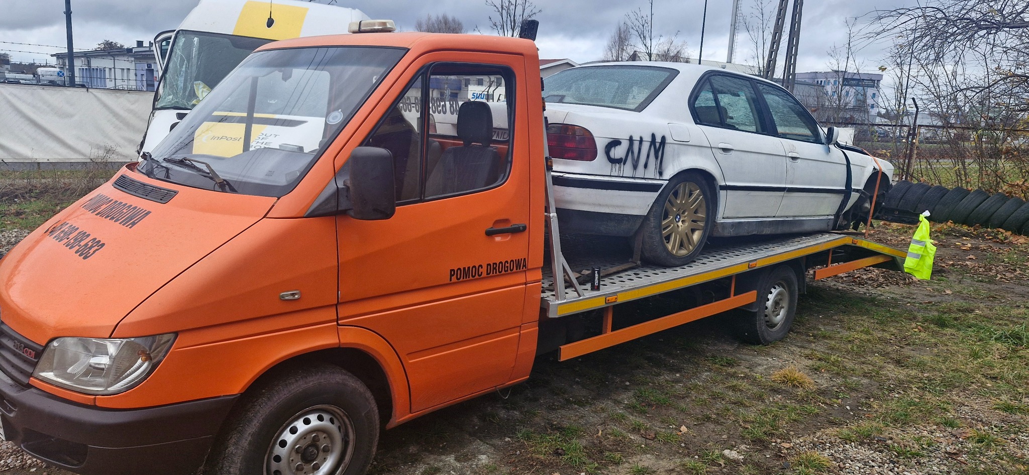 Pomarańczowa laweta z napisem 'Pomoc Drogowa' przewozi uszkodzony, biały samochód z napisem 'CNM' na boku, na tle jesiennego krajobrazu.