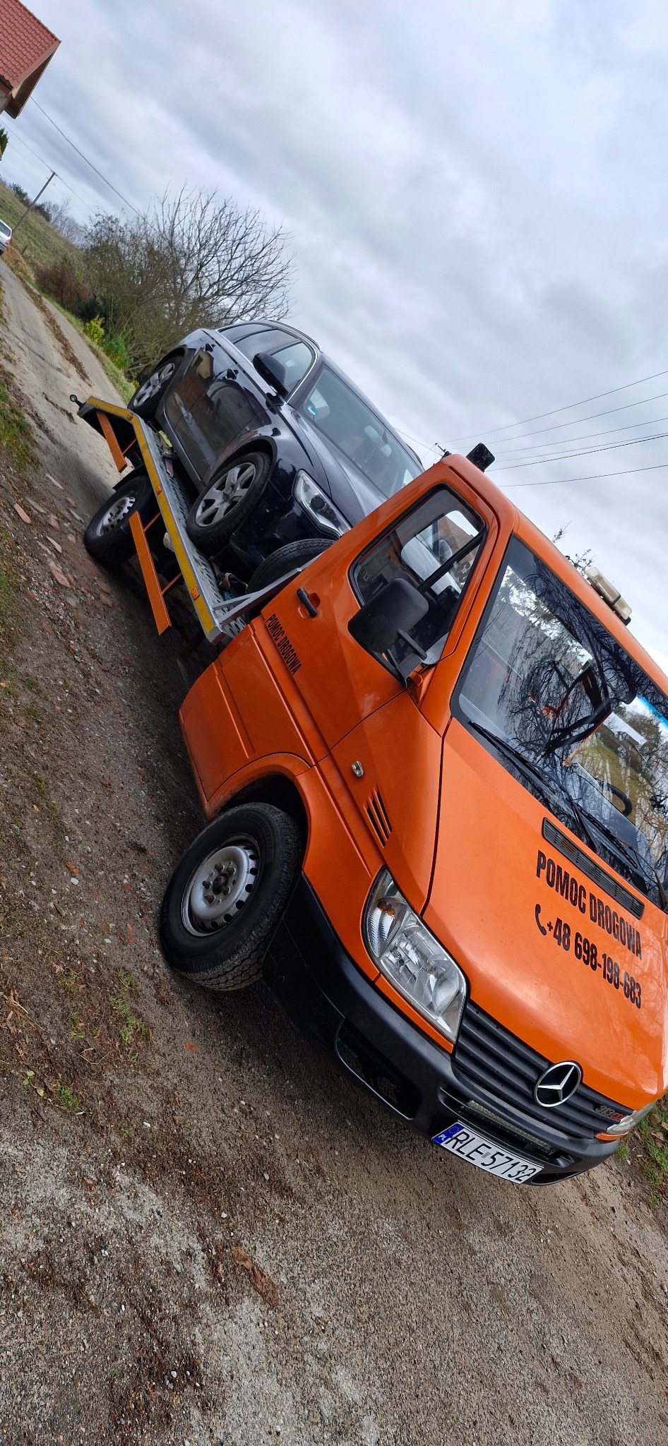 Pomarańczowa laweta z czarnym samochodem osobowym na platformie, widok z boku na tle pochmurnego nieba i polnej drogi.