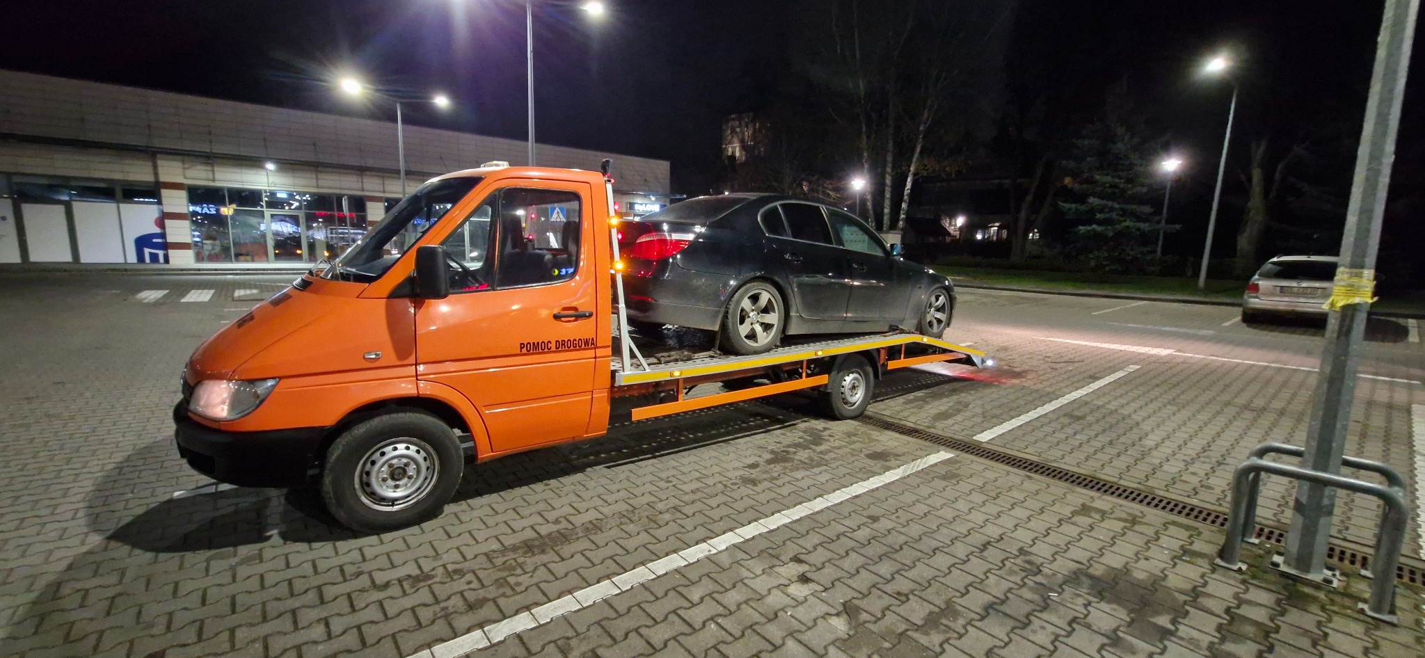 Pomarańczowa laweta z czarnym samochodem osobowym na platformie, stojąca na parkingu przed budynkiem sklepu nocą, widoczne światła ostrzegawcze.