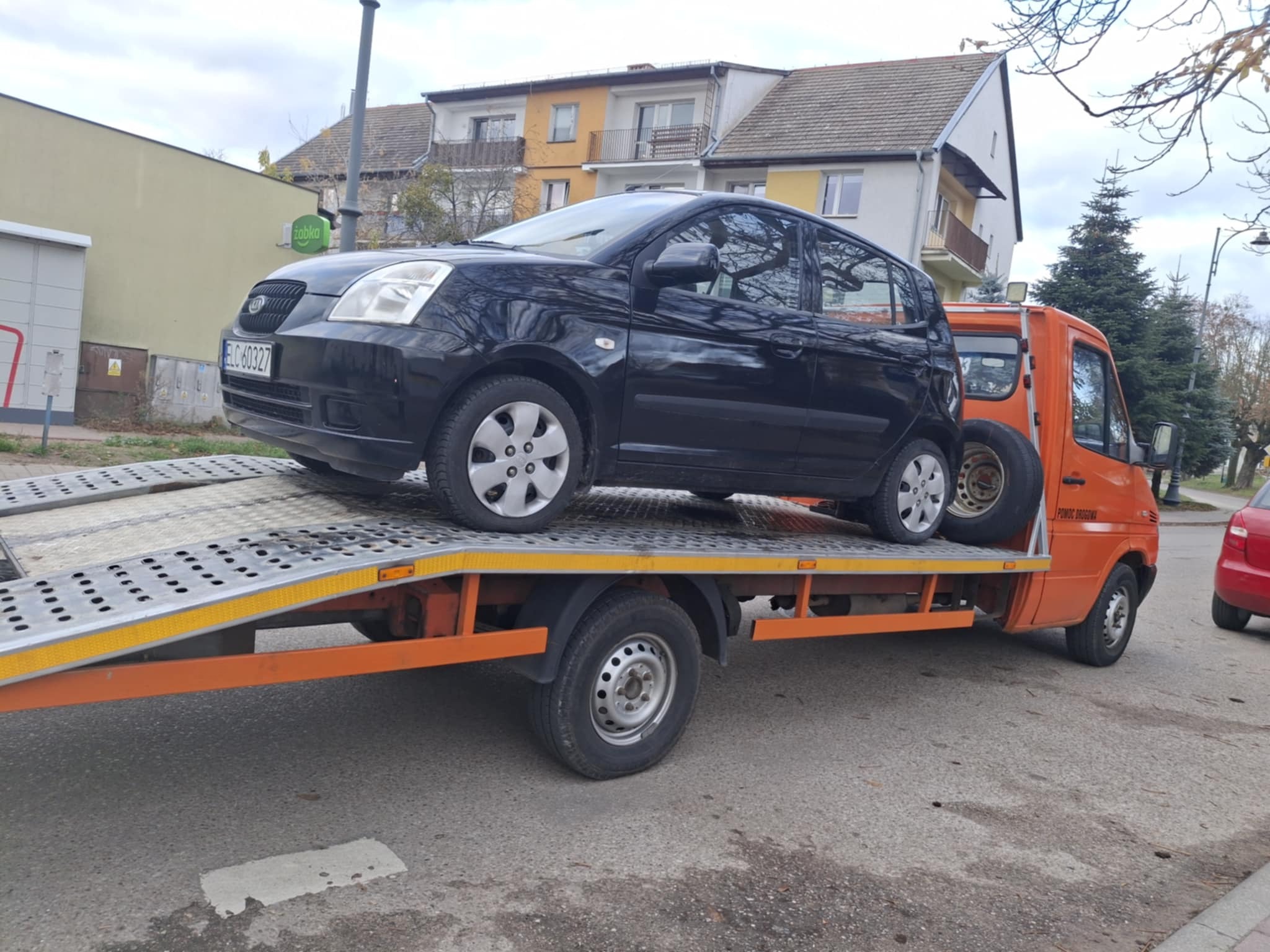 Czarny samochód osobowy Kia na pomarańczowej lawecie z napisem 'Pomoc Drogowa' na tle miejskiego krajobrazu z budynkami i drzewami.