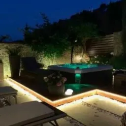 Oświetlona strefa relaksu z hydromasażem na zewnątrz, widok nocą, otoczona dekoracyjnym oświetleniem LED i kamienną ścieżką.