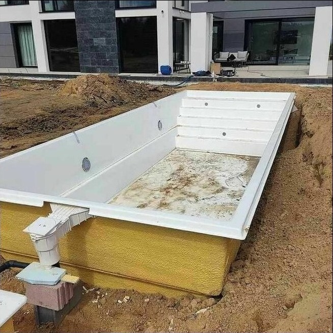 Wkopany w ziemię, pusty basen z białym wnętrzem i żółtą izolacją, widoczne schody i instalacja odpływowa, w tle dom z dużymi oknami.